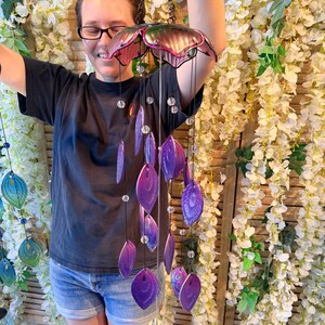A woman is smiling and holding a colorful wind chime with purple and pink beads hanging from it.