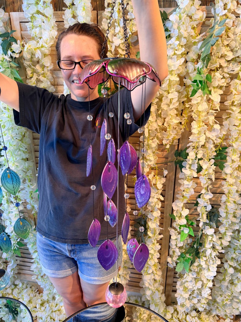 A woman is smiling and holding a colorful wind chime with purple and pink beads hanging from it.