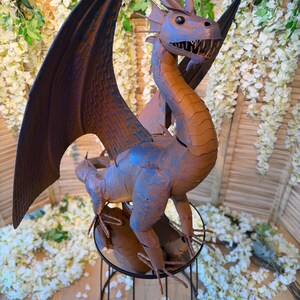 A large metal dragon statue with outstretched wings stands on a metal stand, surrounded by white flowers and greenery.