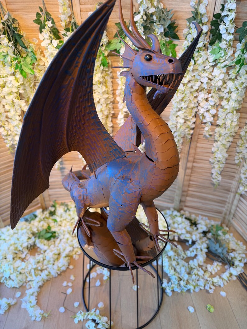 A large metal dragon statue with outstretched wings stands on a metal stand, surrounded by white flowers and greenery.