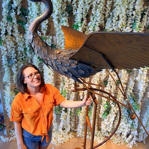 A woman in an orange shirt stands next to a large metal sculpture of a bird, which appears to be a windmill-like structure.