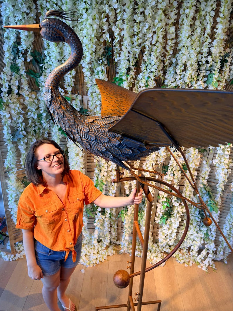 A woman in an orange shirt stands next to a large metal sculpture of a bird, which appears to be a windmill-like structure.