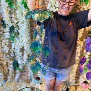 A woman is standing in front of a floral backdrop, holding a colorful glass wind chime with blue and green glass elements.