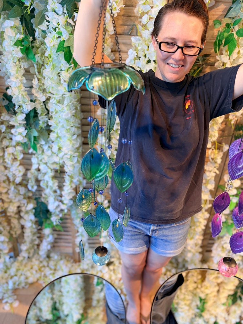 A woman is standing in front of a floral backdrop, holding a colorful glass wind chime with blue and green glass elements.