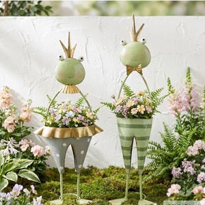 May include: Two whimsical frog garden statues. One frog wears a gold crown and a polka-dot skirt, the other wears a bow tie and striped pants. Both hold baskets filled with colorful flowers. The statues are green, gold, and gray.