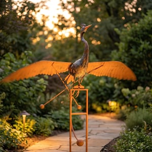 A large metal bird sculpture with orange wings stands on a stone path in a lush garden, surrounded by greenery and illuminated by warm lighting.