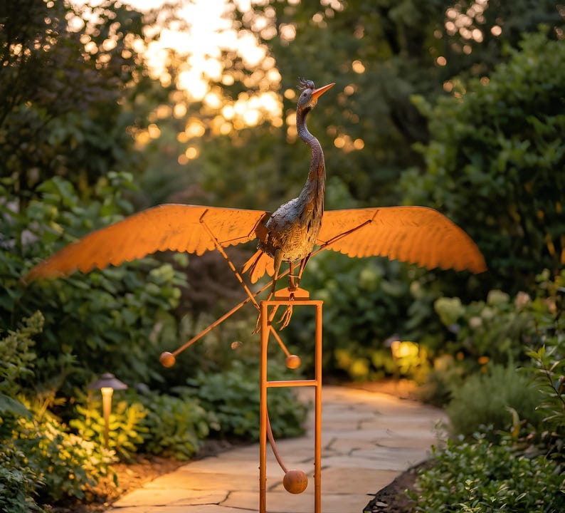 A large metal bird sculpture with orange wings stands on a stone path in a lush garden, surrounded by greenery and illuminated by warm lighting.