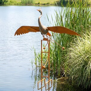 A large metal bird sculpture with orange wings and a long neck stands on a metal stand in a body of water, surrounded by tall grass.