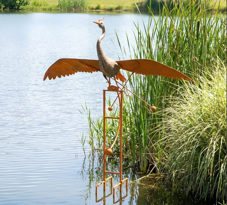 A large metal bird sculpture with orange wings and a long neck stands on a metal stand in a body of water, surrounded by tall grass.