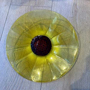 May include: A circular glass plate featuring a bright yellow translucent rim with a textured, petal-like design radiating from the center. The center of the plate is a dark, deep brown circular element with a raised, dotted, seed-like texture.