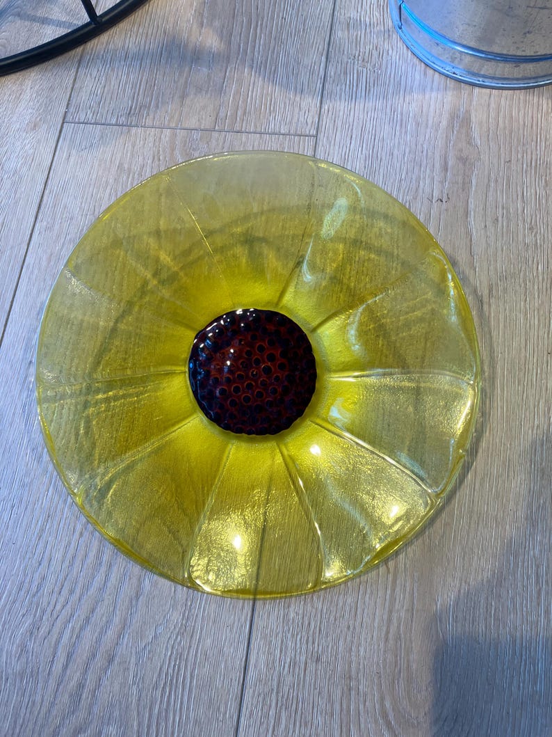 May include: A circular glass plate featuring a bright yellow translucent rim with a textured, petal-like design radiating from the center. The center of the plate is a dark, deep brown circular element with a raised, dotted, seed-like texture.