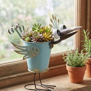 A blue metal bird figurine holding a potted plant with various succulents and other greenery, placed on a windowsill.
