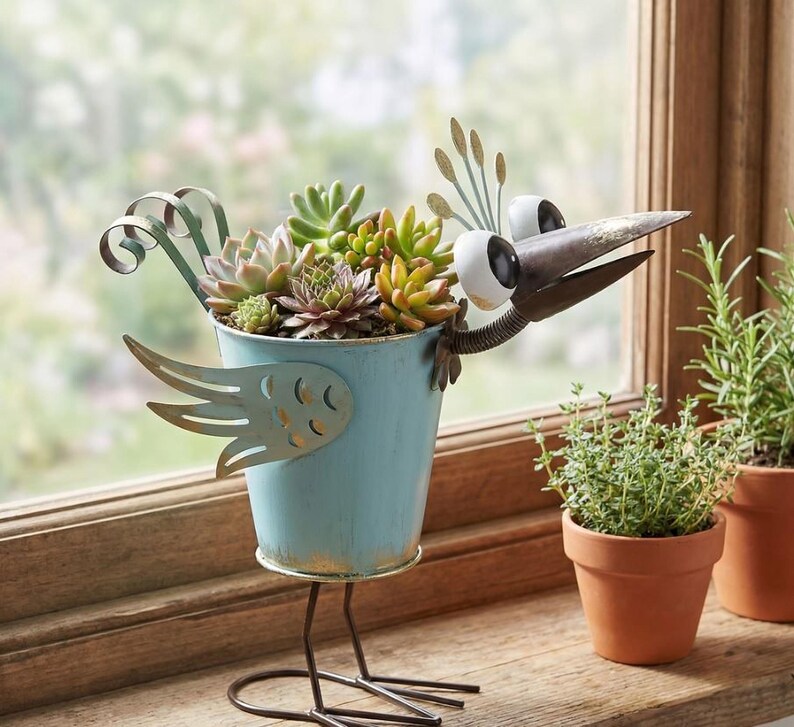 A blue metal bird figurine holding a potted plant with various succulents and other greenery, placed on a windowsill.