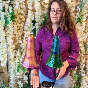 A woman in a purple shirt stands in front of a floral backdrop, holding two colorful glass lamps.