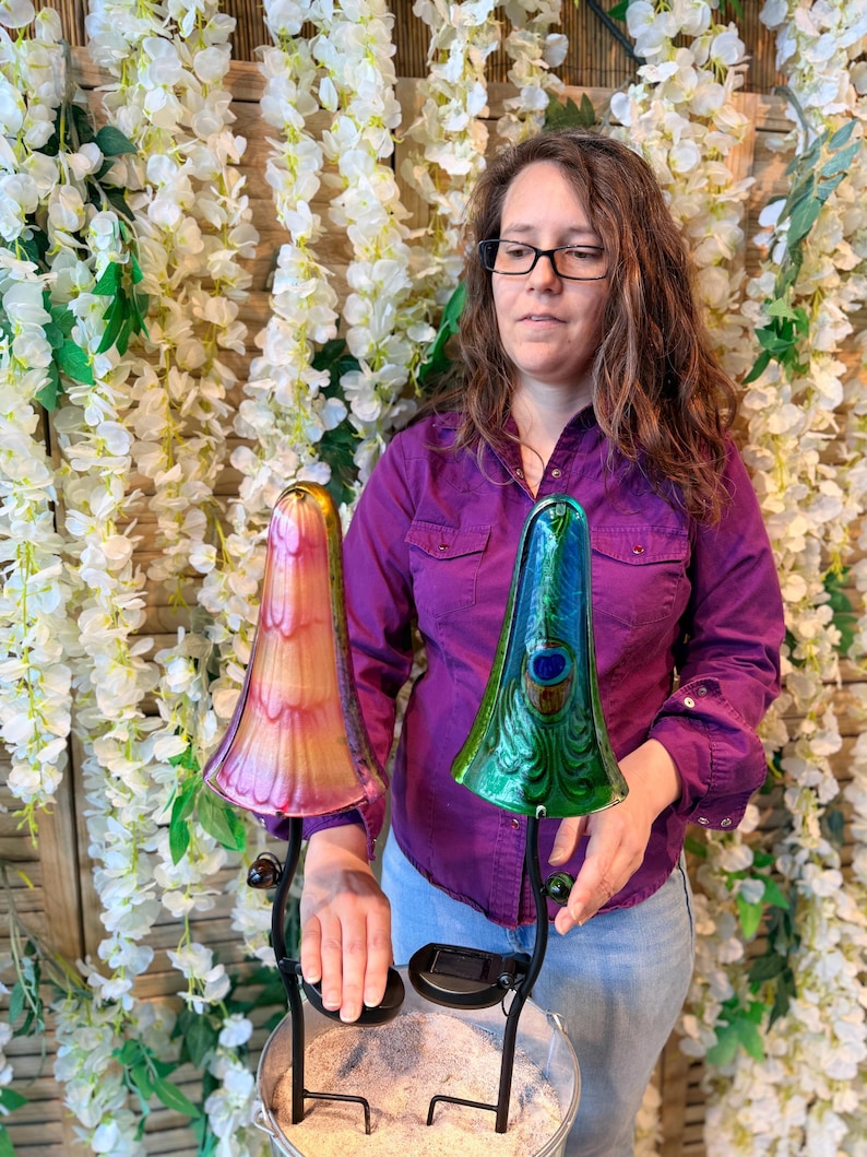 A woman in a purple shirt stands in front of a floral backdrop, holding two colorful glass lamps.