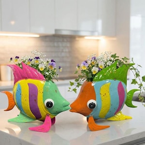 Può includere: Due fioriere a forma di pesce in metallo colorato con vivaci tonalità di rosa, giallo, verde e blu. Ogni pesce ha un occhio grande e contiene una piccola composizione di fiori e verde, creando un accento decorativo stravagante.