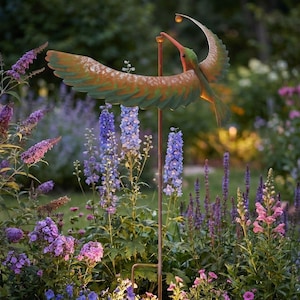 Puede incluir: Una estaca de jardín de metal con forma de colibrí con alas extendidas, pintadas en tonos naranja y verde. La estaca tiene una pequeña luz en la parte superior y está situada en un jardín de flores moradas y rosas.
