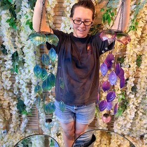 A woman is standing in front of a floral backdrop, holding up two colorful wind chimes with shells and beads hanging from them.