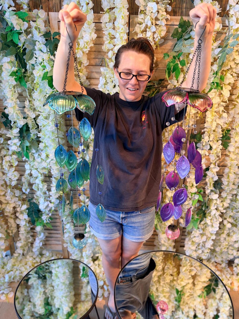 A woman is standing in front of a floral backdrop, holding up two colorful wind chimes with shells and beads hanging from them.