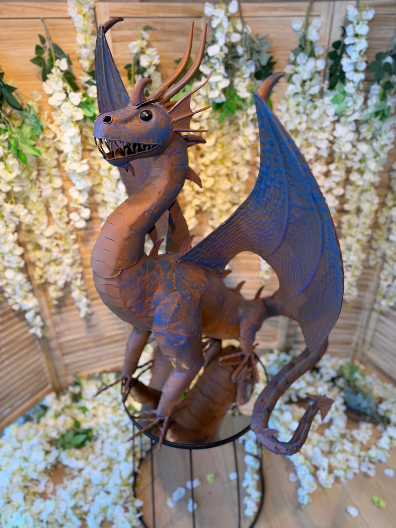 A large, detailed dragon sculpture with blue and brown hues stands prominently in the center of the image, surrounded by white flowers and greenery.