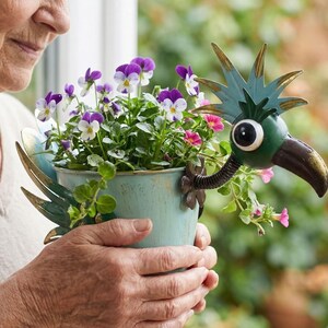 An elderly woman holds a blue pot filled with purple and white flowers, which are decorated with a bird-like metal sculpture.