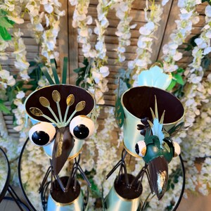 May include: Two whimsical bird-shaped planters. The metal sculptures feature large, round eyes, long beaks, and decorative details in gold and green. The planters are set on a glass table with a floral background.
