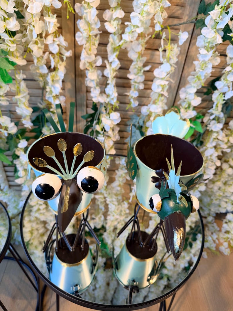 May include: Two whimsical bird-shaped planters. The metal sculptures feature large, round eyes, long beaks, and decorative details in gold and green. The planters are set on a glass table with a floral background.