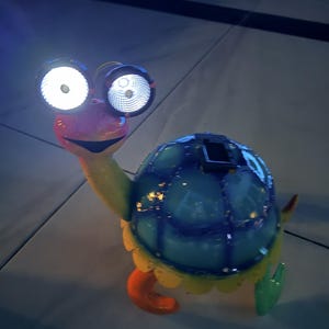 May include: A colorful turtle-shaped solar-powered garden light. The turtle has large, bright, round eyes and a blue shell. Its legs are orange and green, and the edges of the shell are yellow. The light is designed to illuminate outdoor spaces.