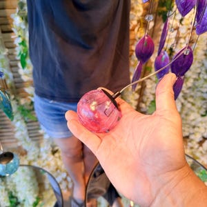 A person's hand holding a small, round, pink object, possibly a glass or a small figurine, with a string attached to it. The background is filled with various colorful objects, including purple and blue items, creating a vibrant and lively atmosphere.