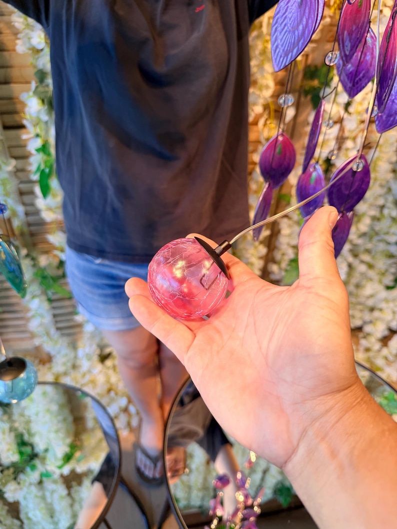 A person's hand holding a small, round, pink object, possibly a glass or a small figurine, with a string attached to it. The background is filled with various colorful objects, including purple and blue items, creating a vibrant and lively atmosphere.