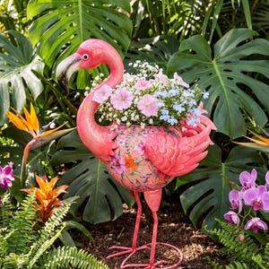 Puede incluir: Una maceta con forma de flamenco rosa que presenta un estampado floral en el cuerpo. La maceta está llena de un ramo de pequeñas flores rosas, blancas y azul claro. El flamenco tiene un cuello largo, un pico curvado con punta negra y patas finas.
