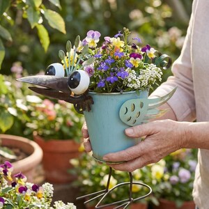 A person is holding a blue metal bird planter filled with colorful flowers, including purple, yellow, and white blooms.