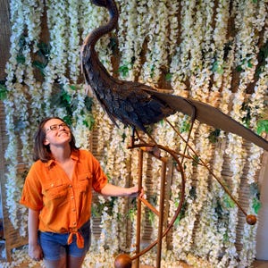 A woman in an orange shirt stands next to a large metal sculpture of a bird, which appears to be a large metal bird perched on a stand.