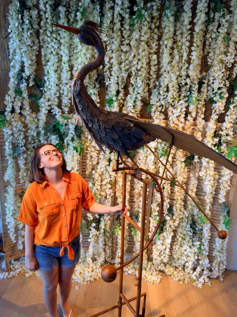 A woman in an orange shirt stands next to a large metal sculpture of a bird, which appears to be a large metal bird perched on a stand.