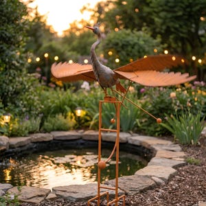 a garden scene with a metal bird sculpture perched on a metal stand, which is situated near a small pond. The bird sculpture has orange wings and a long neck, and it appears to be in motion, as if it is flying. The garden is filled with various plants and flowers, creating a serene and picturesque setting.