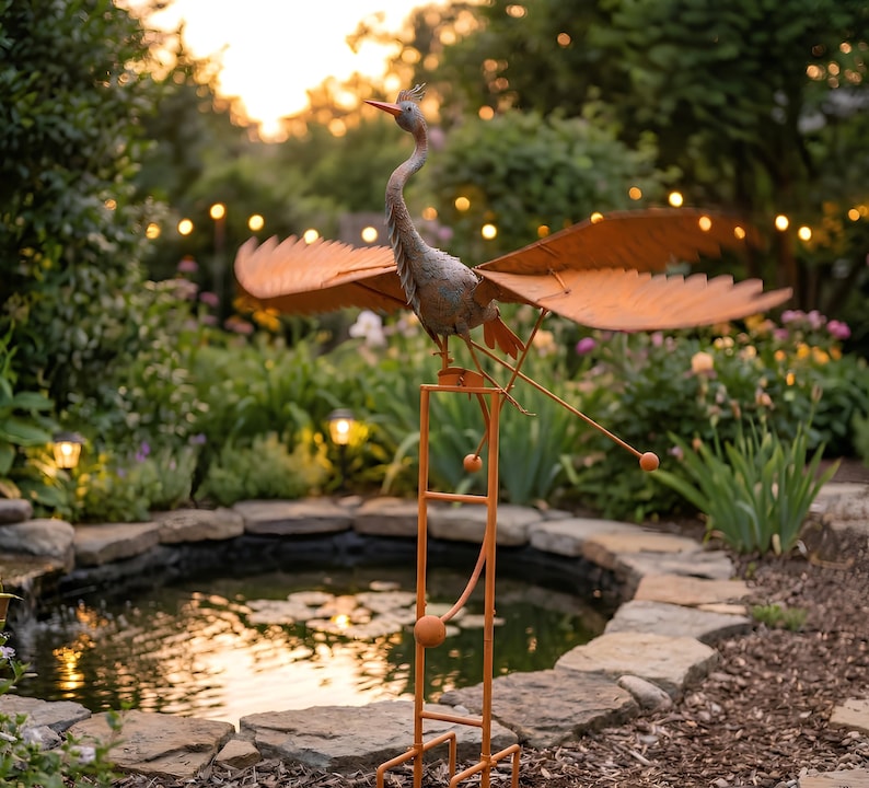 a garden scene with a metal bird sculpture perched on a metal stand, which is situated near a small pond. The bird sculpture has orange wings and a long neck, and it appears to be in motion, as if it is flying. The garden is filled with various plants and flowers, creating a serene and picturesque setting.