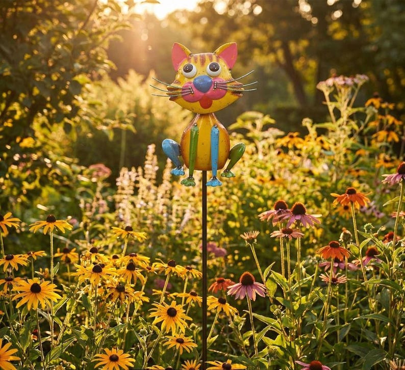 A colorful metal cat decoration stands in a vibrant garden filled with yellow flowers and lush greenery.