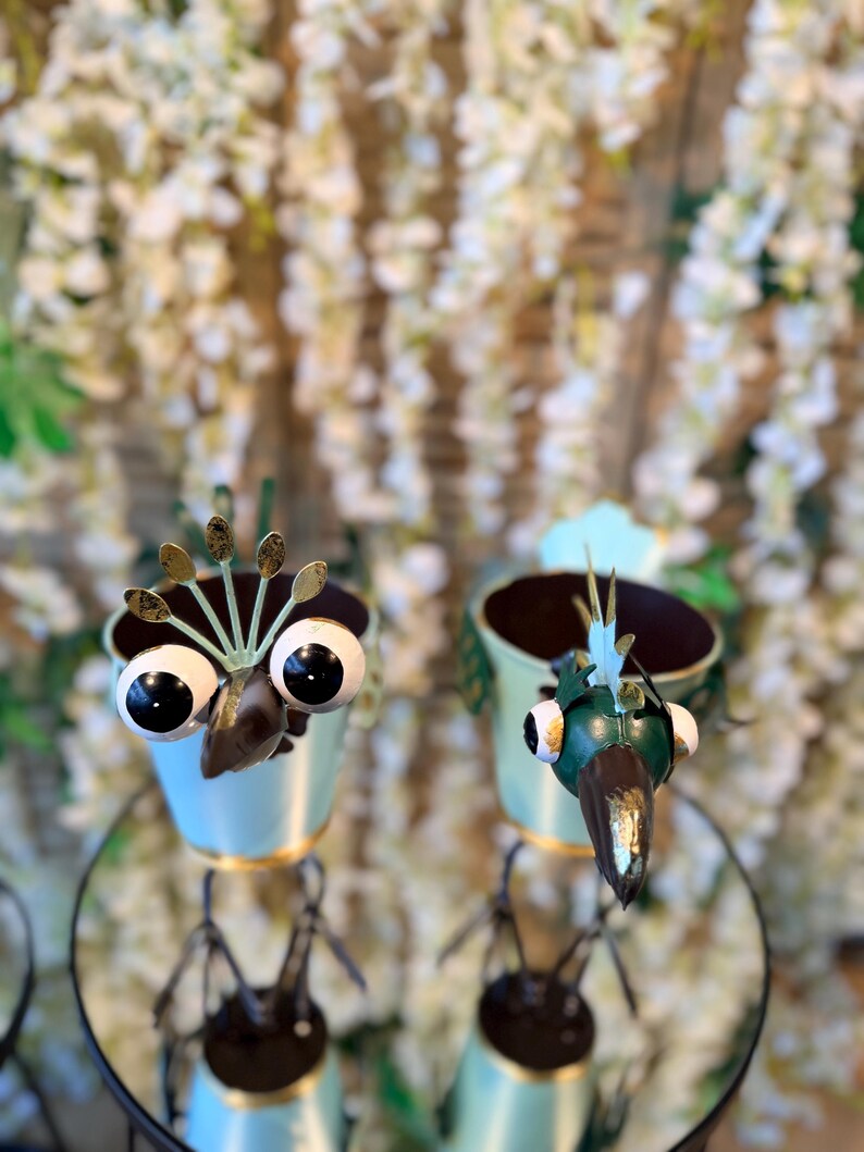 May include: Two whimsical bird-shaped planters. Each planter has a light blue body with gold trim, large googly eyes, and a brown beak. The planters are made of metal and have a dark brown interior.