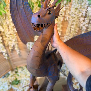 A person's hand is holding a large, detailed dragon sculpture made of metal or clay. The dragon has a blue and brown color scheme and is positioned in front of a backdrop of white flowers.
