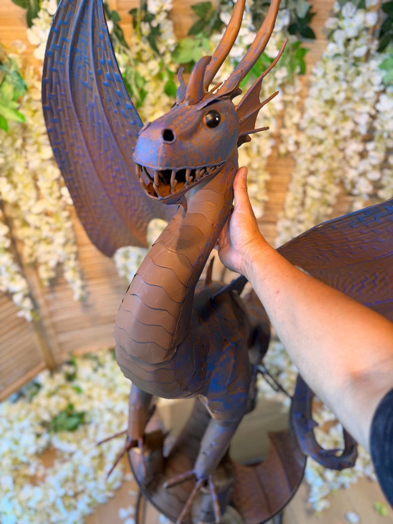 A person's hand is holding a large, detailed dragon sculpture made of metal or clay. The dragon has a blue and brown color scheme and is positioned in front of a backdrop of white flowers.