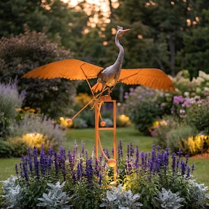 A large metal bird sculpture with orange wings perched on a metal stand, surrounded by a colorful garden with purple flowers and lush greenery.