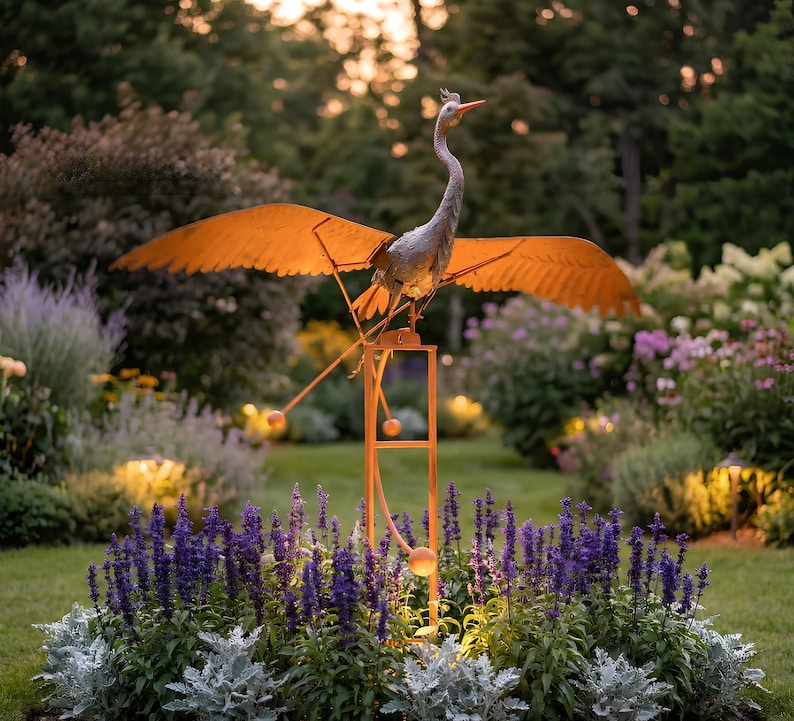 A large metal bird sculpture with orange wings perched on a metal stand, surrounded by a colorful garden with purple flowers and lush greenery.