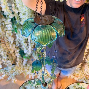A person is holding a green glass wind chime with blue and green glass beads, standing in front of a floral backdrop.