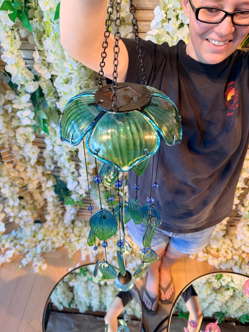 A person is holding a green glass wind chime with blue and green glass beads, standing in front of a floral backdrop.