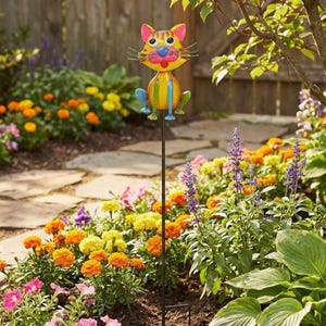 A colorful garden decoration resembling a cat, with a metal pole in the foreground and a variety of flowers and plants in the background.