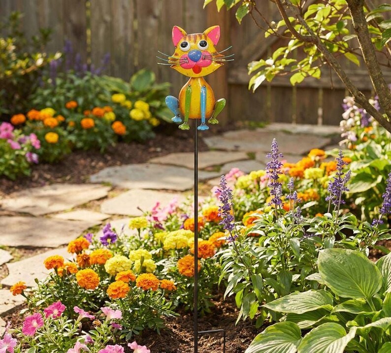 A colorful garden decoration resembling a cat, with a metal pole in the foreground and a variety of flowers and plants in the background.