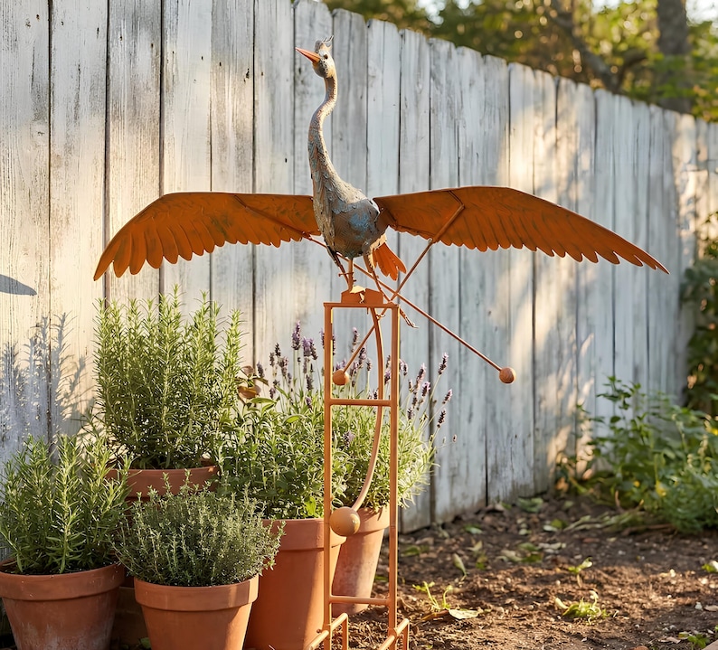 A large metal bird sculpture with orange wings perched on a metal stand, surrounded by potted plants in a garden setting.