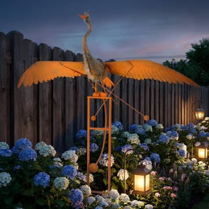 A large orange bird sculpture with outstretched wings stands on a metal stand in a garden, surrounded by colorful flowers and lit lanterns.