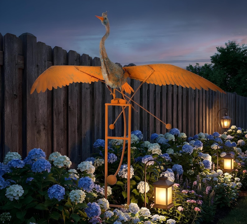 A large orange bird sculpture with outstretched wings stands on a metal stand in a garden, surrounded by colorful flowers and lit lanterns.