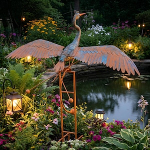 A large metal sculpture of a heron stands on a metal stand in a garden, surrounded by colorful flowers and plants. The heron's wings are spread wide, and it is perched on a ladder, creating a whimsical and artistic scene.
